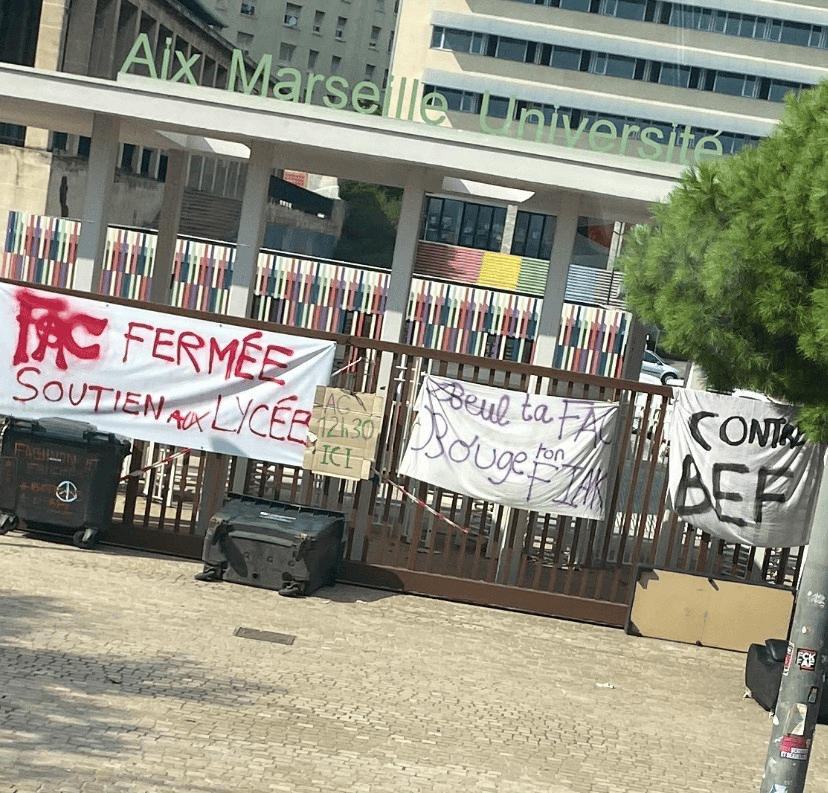 Aix Marseille Üniversitesi önünde protesto pankartları asılmış, çöp kutuları görülebiliyor.